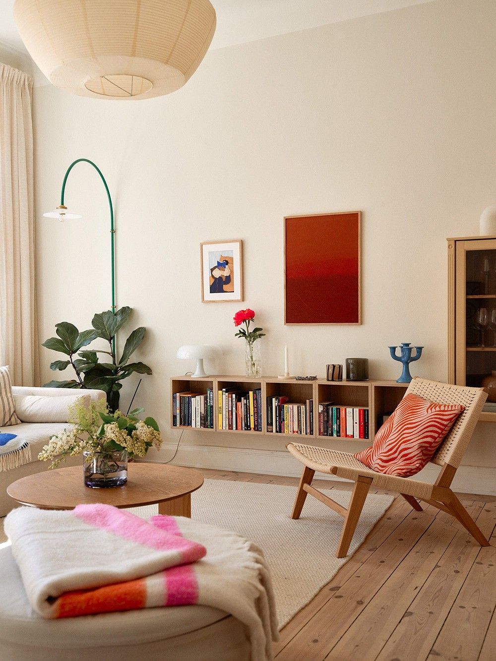A sunlit apartment corner with a low chair, a small side table stacked with books, and a wall-mounted reading light angled toward the seat