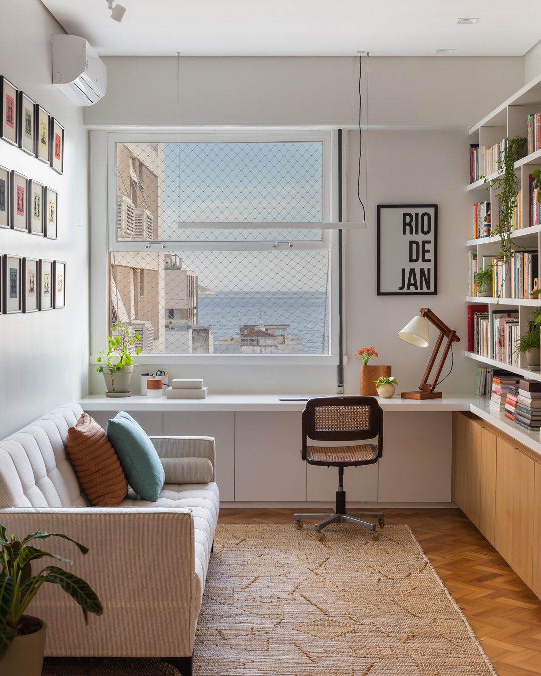 A tidy corner workspace in a small apartment bedroom, with a folding desk, monitor riser, and a single architectural lamp casting warm directional light over a keyboard